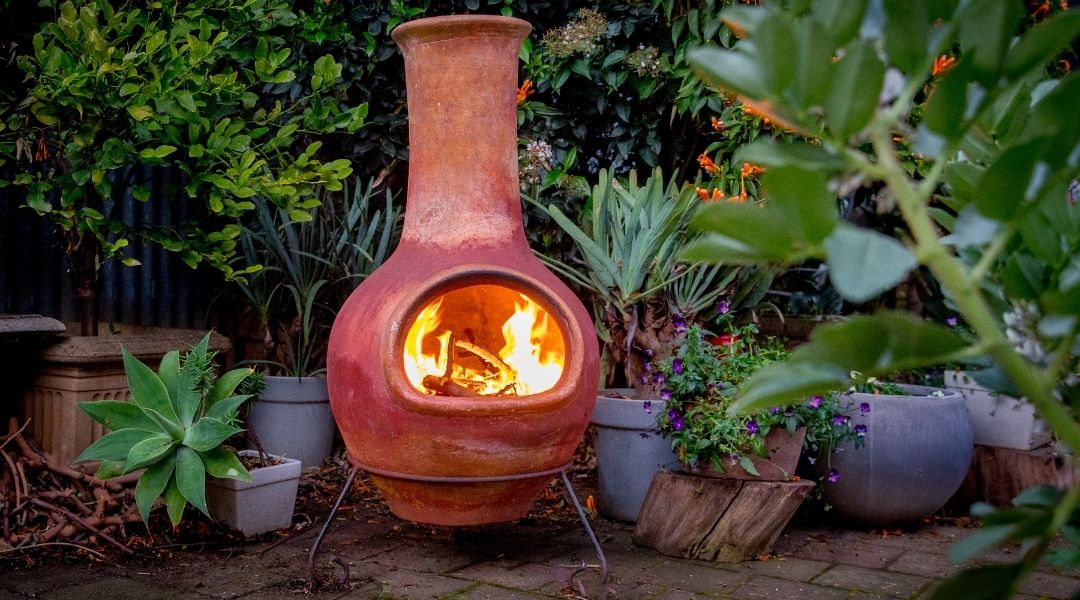 A clay chiminea on metal stand with a fire going surrounded by plants